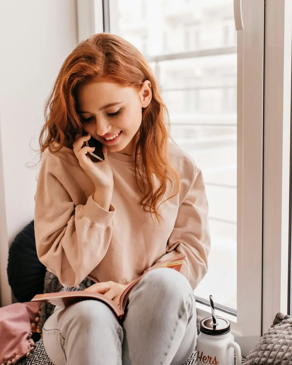 Une jeune femme assise près d'une fenêtre, un carnet sur les genoux, parle au téléphone en souriant